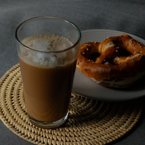 A big glas with black tea and plantbased milk. In the background a prezel on a plate.